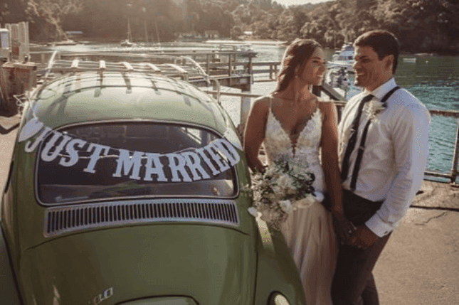 Bride and groom standing by a green car with 'Just Married' sign, near a waterfront.